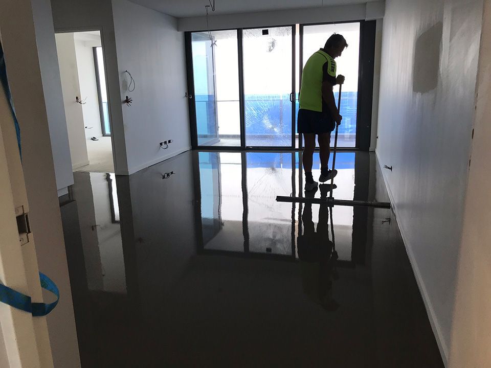 Person smoothing a wet, dark gray floor in a mostly empty room with floor-to-ceiling windows.