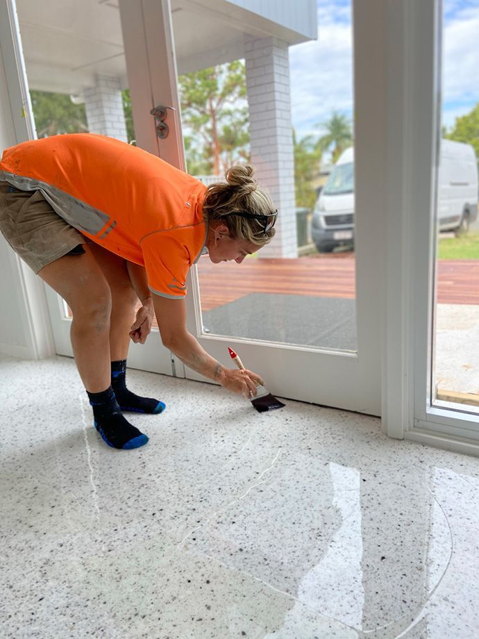 Woman painting a white door frame on a speckled floor, wearing an orange shirt and shorts.