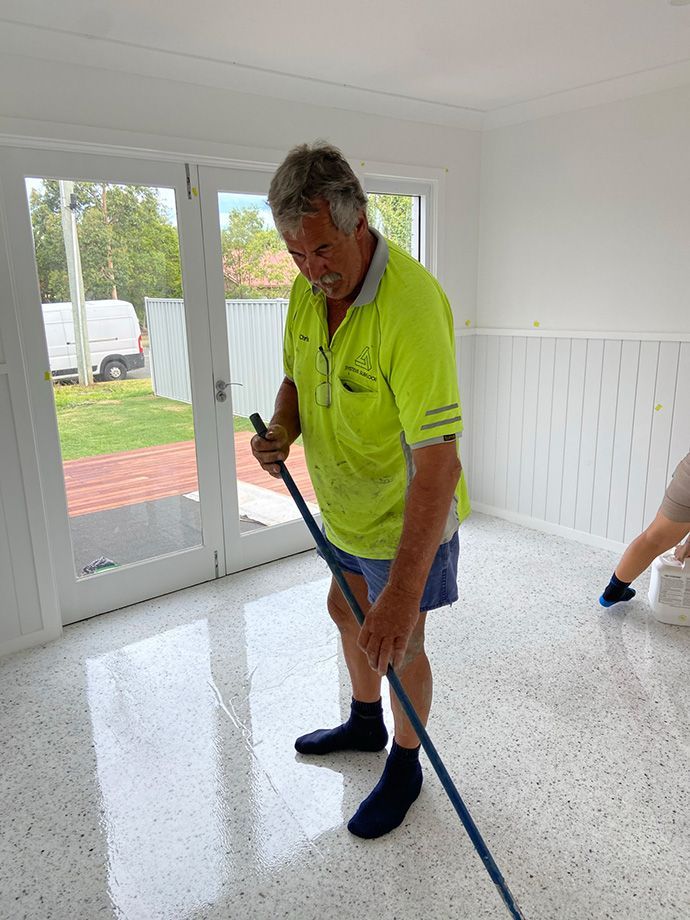 Man mopping a shiny, speckled floor in a bright room with large windows.
