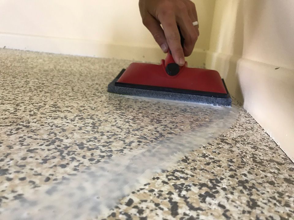 Hand applying a clear sealant with a red tool to a speckled floor near a wall.