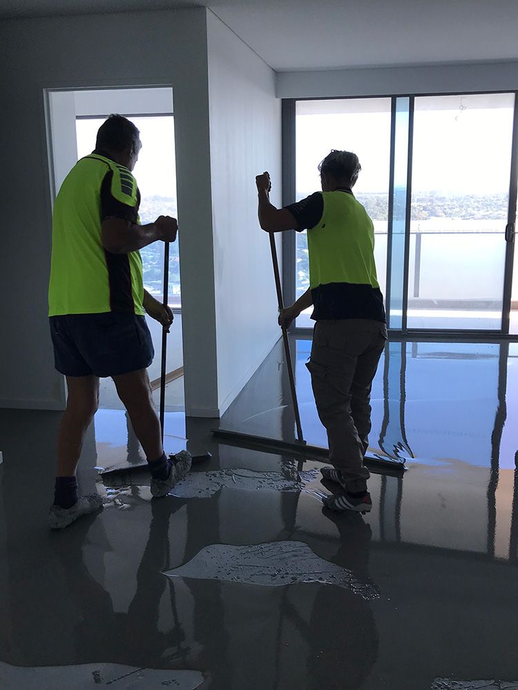 Two workers in safety vests cleaning a floor with squeegees in a room with a waterfront view.