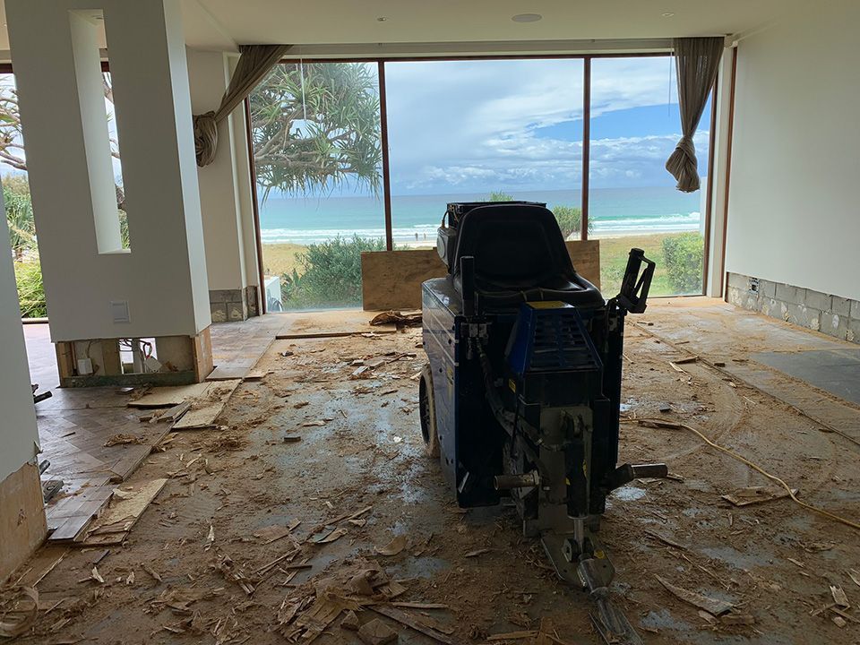 Room under renovation with ocean view. Floor scraper in the middle, debris scattered, large windows overlooking the beach.