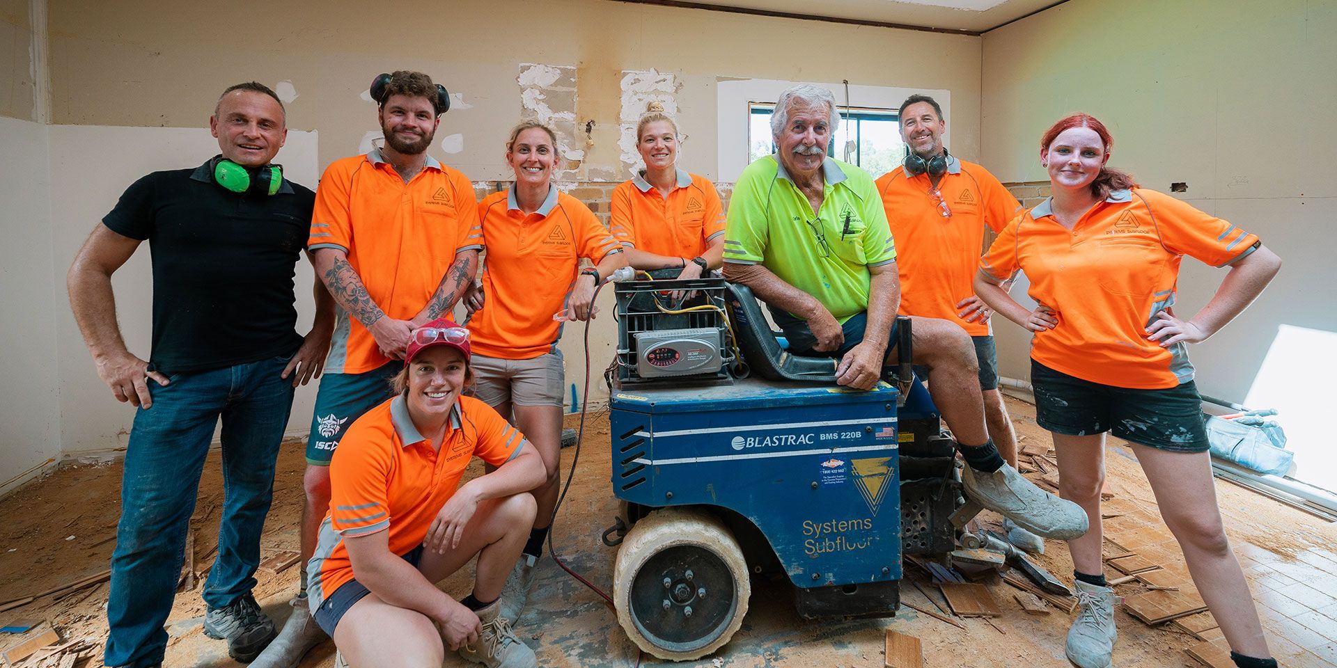 A construction crew poses in a room with a machine. They wear work uniforms, and the room appears to be under renovation.
