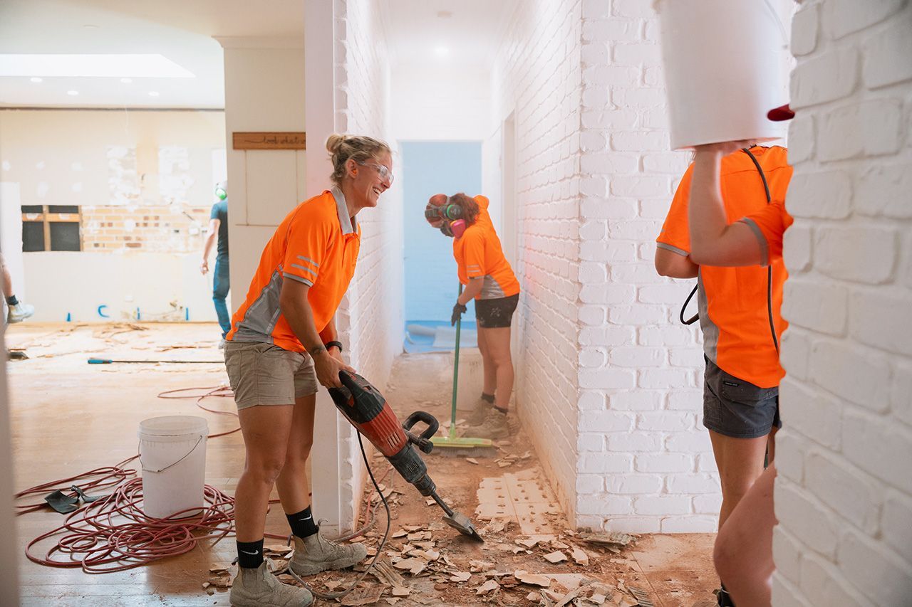 Three people wearing orange shirts work on a construction site. One uses a jackhammer.