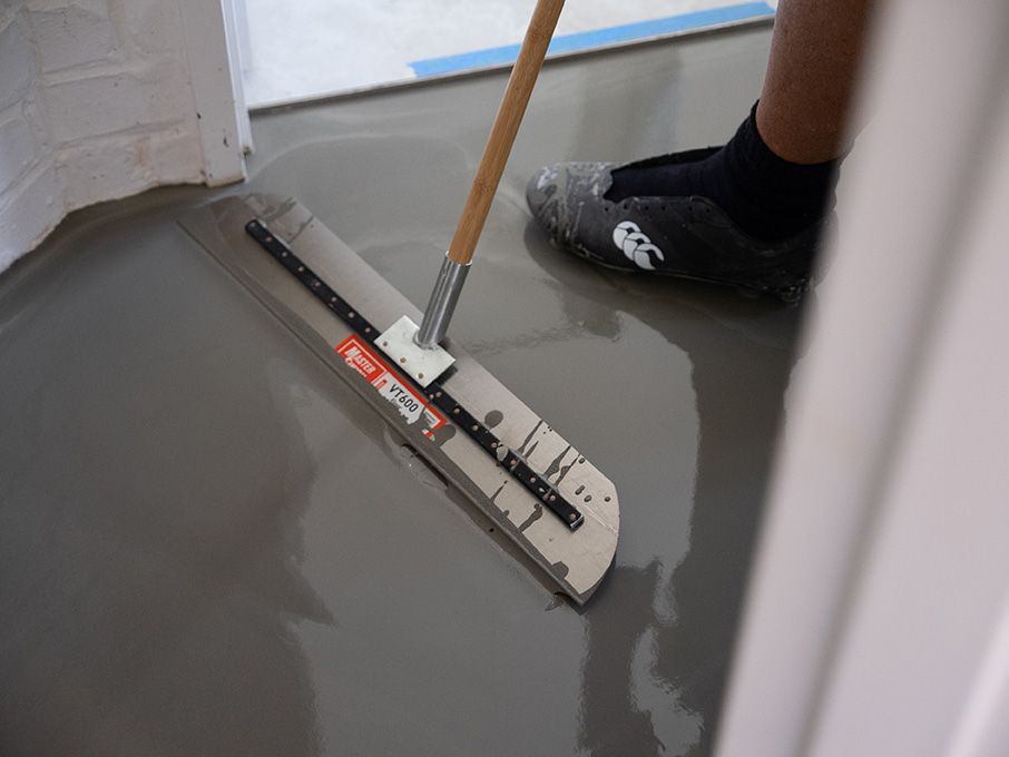 Person smoothing wet, gray floor with a long trowel in a room during construction.