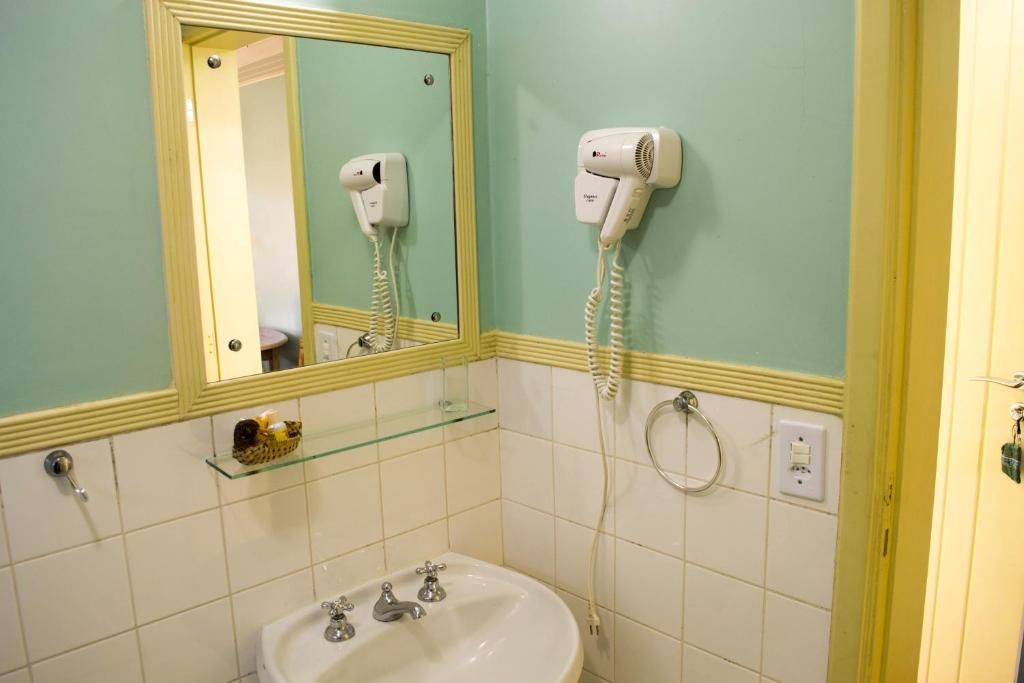 Uma pia de banheiro com prateleira de vidro, espelho e secador de cabelo fixado na parede, em contraste com paredes verde-claras e backsplash de azulejos brancos.