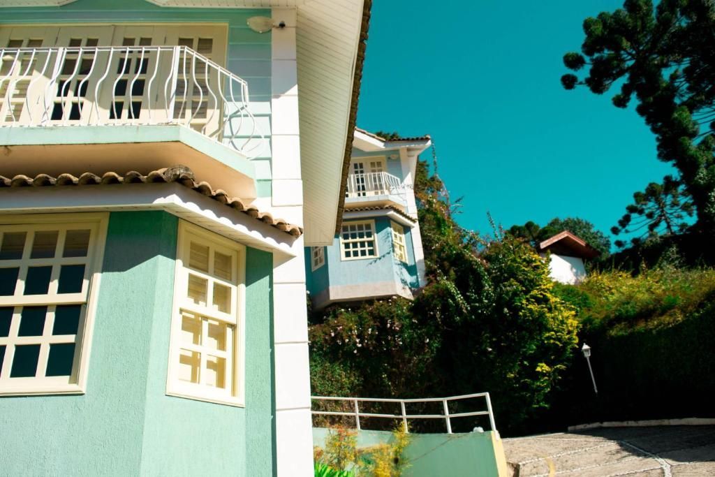 Duas casas de vários andares, em tom azul claro, erguem-se em uma área pavimentada e inclinada, cercada por árvores e vegetação exuberante, sob um céu límpido.