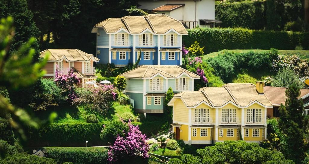 Três casas coloridas — azul, verde e amarela — aninhavam-se em uma paisagem exuberante e montanhosa, repleta de árvores e arbustos floridos.
