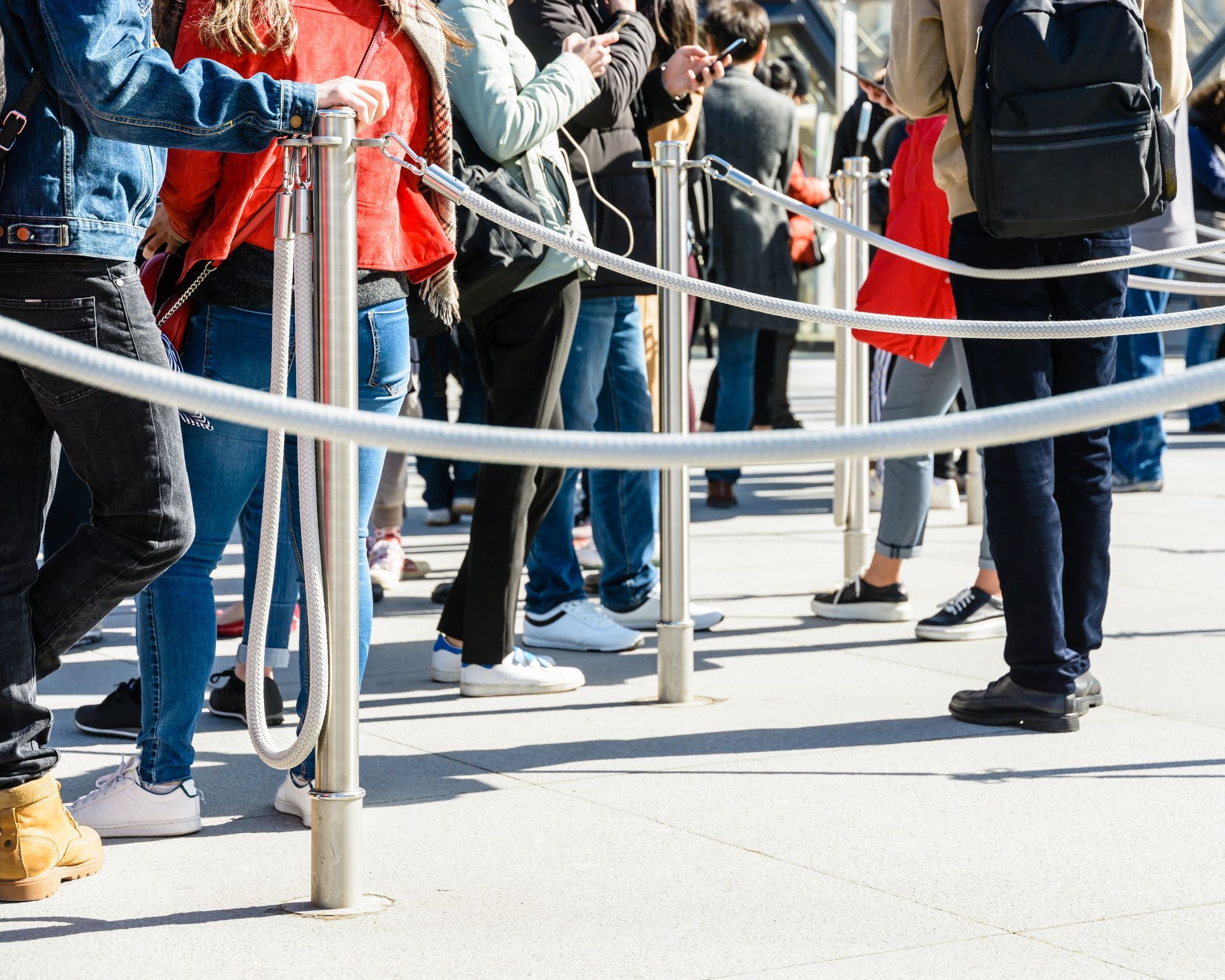 People standing in a line behind metal stanchions outdoors, wearing coats and backpacks