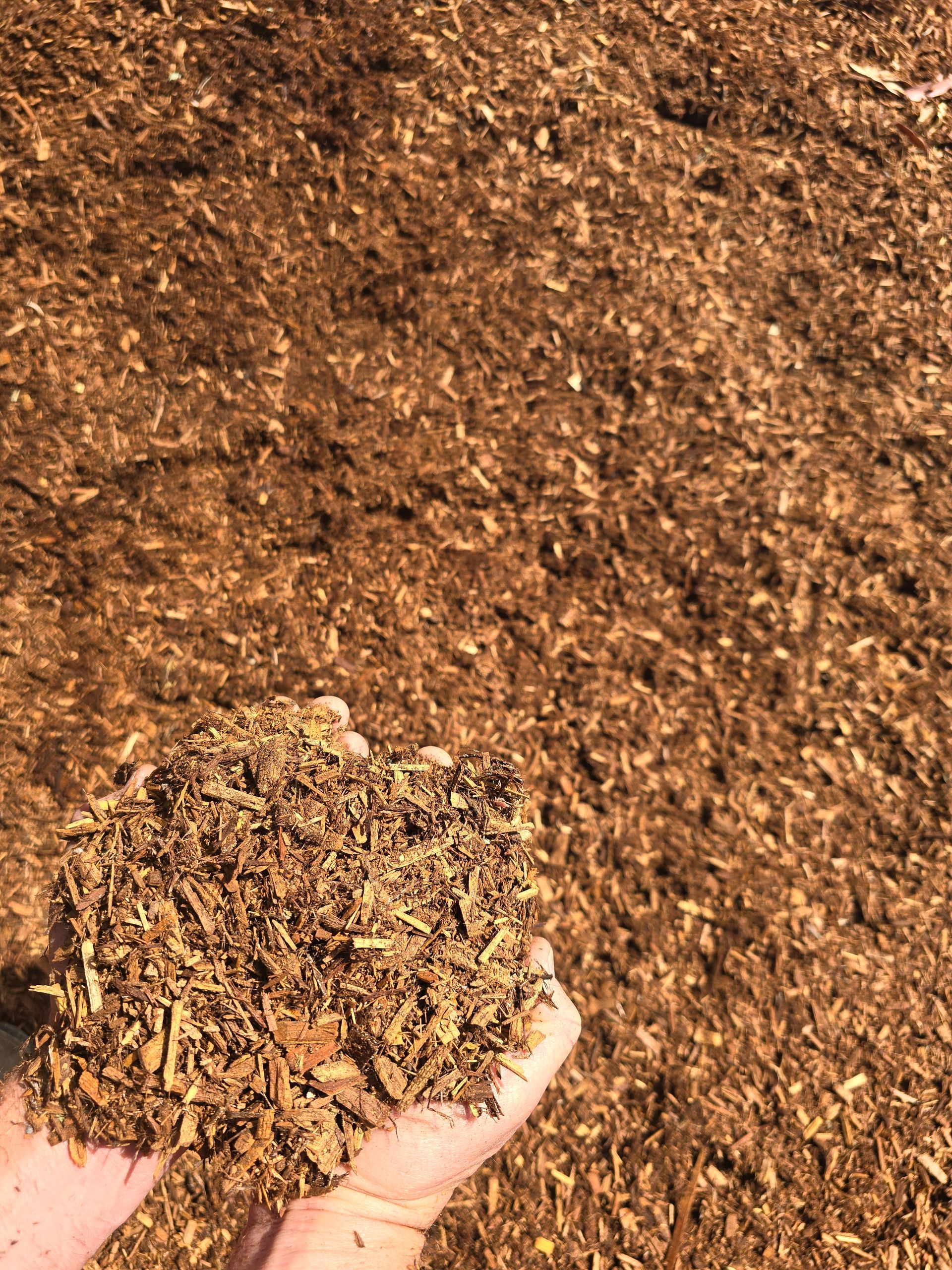 Hand Holding a Pile of Brown Mulch Over a Large Pile of Mulch — BVN Landscape and Concrete Supplies in St Georges Basin, NSW