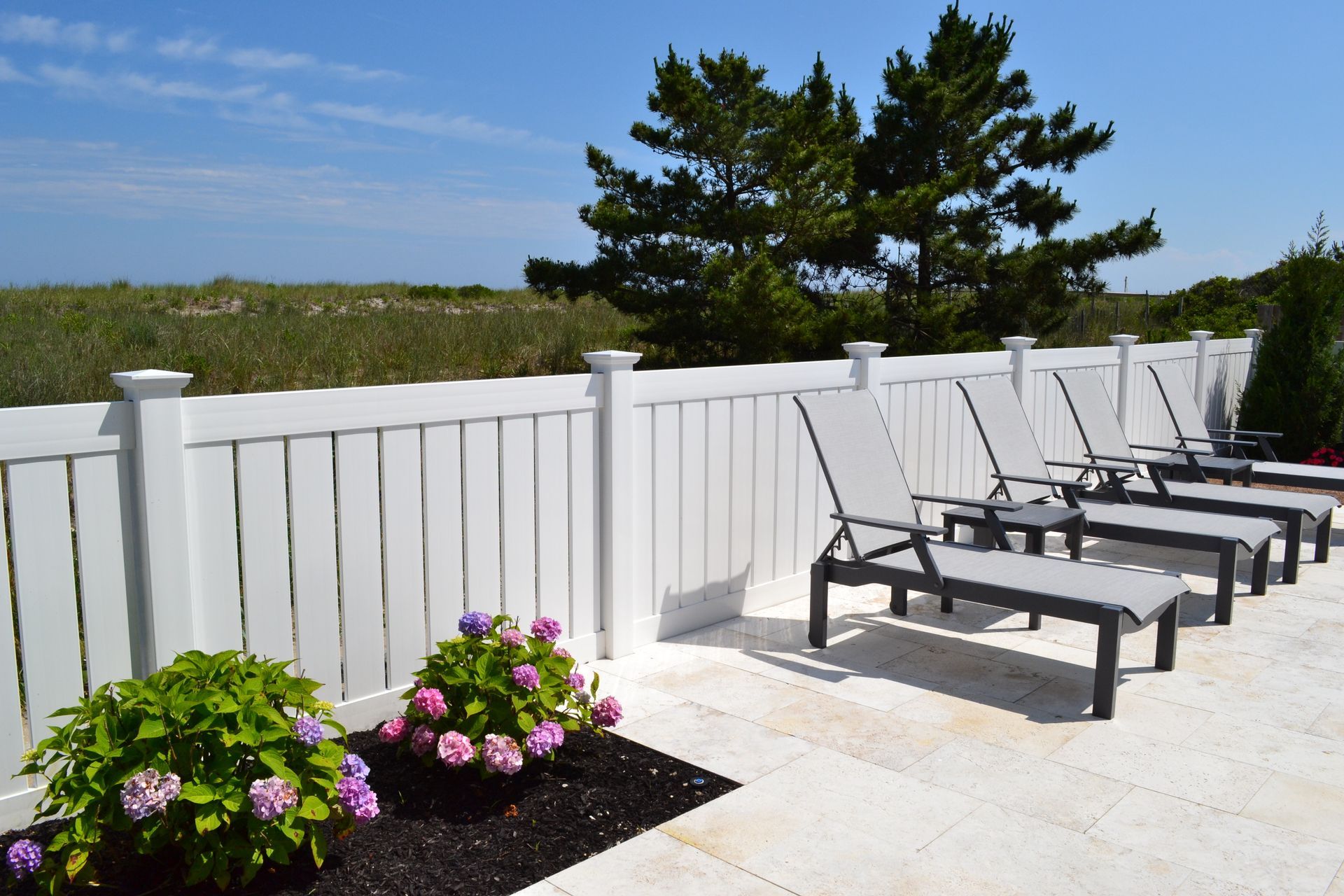 Photograph of an outdoor patio area featuring a row of six black-framed lounge chairs with gray mesh seats positioned against a white wooden fence. Photograph of an outdoor patio area featuring a row of six black-framed lounge chairs with gray mesh seats positioned against a white wooden fence.