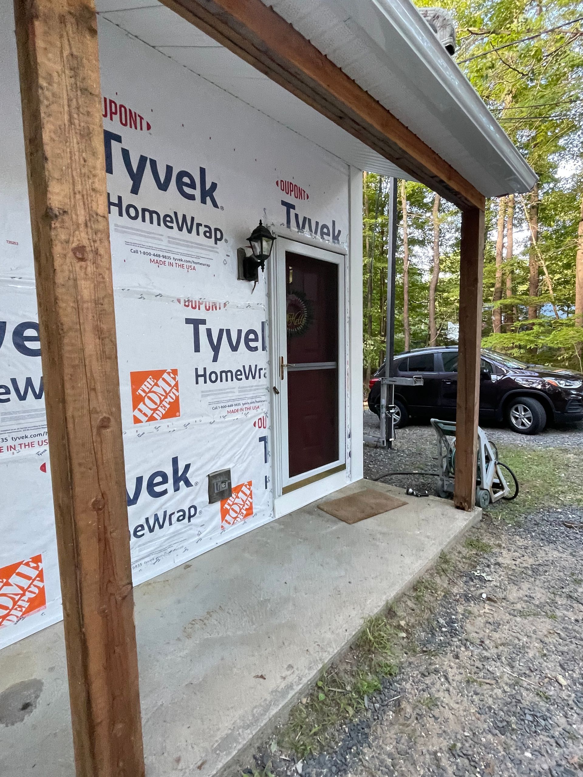 Partially wrapped house exterior with Tyvek HomeWrap, front door, porch, and parked car beside the driveway
