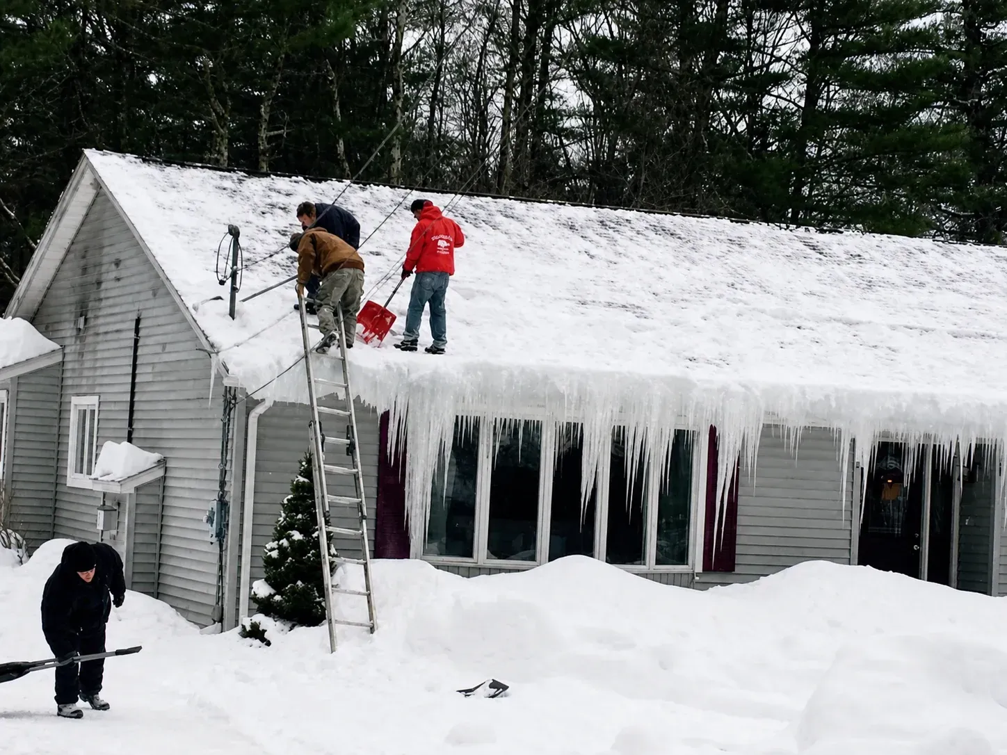 Workers on a snowy roof clearing ice and snow from a house with heavy icicles.
