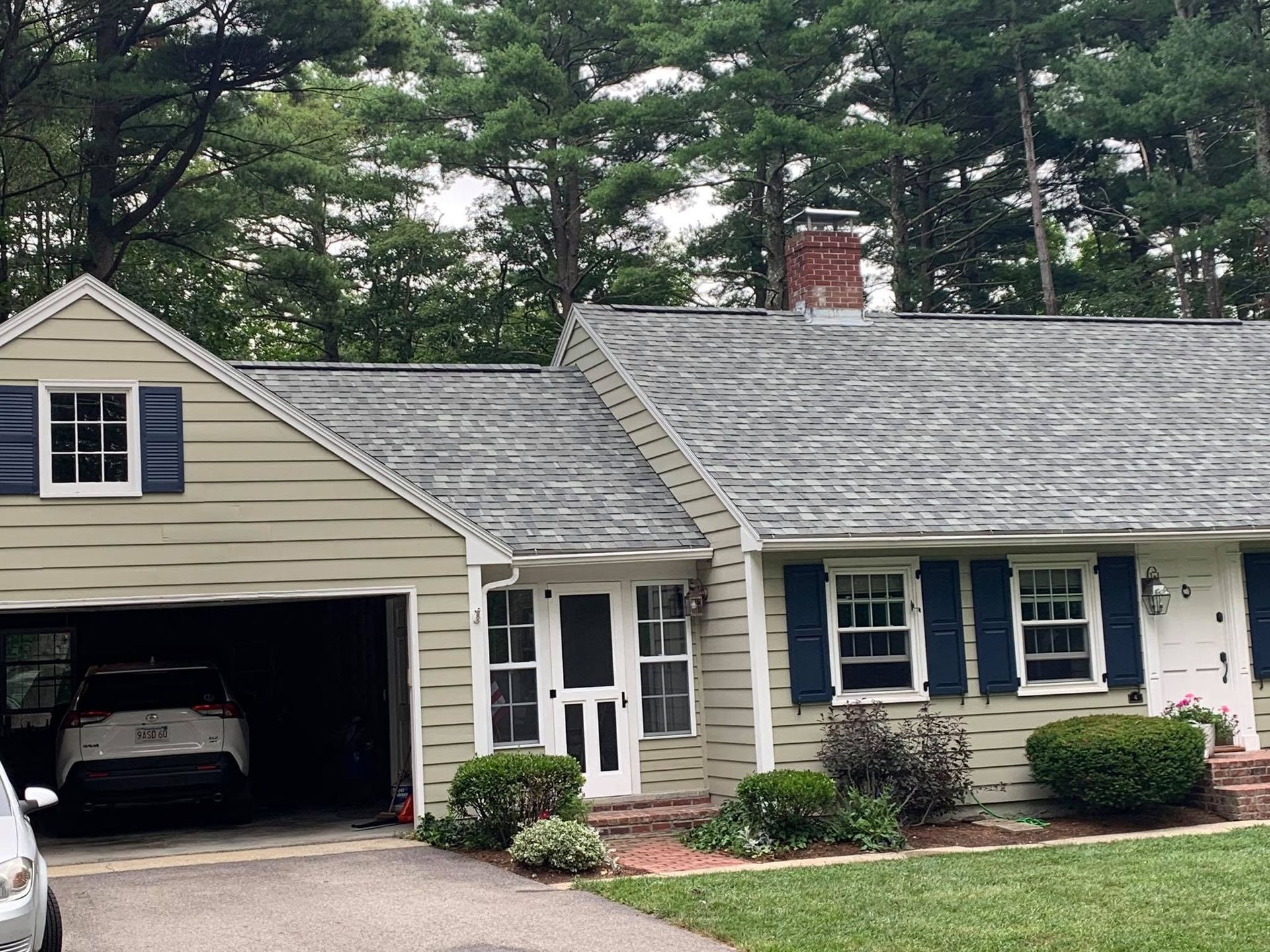 Single-story beige house with open garage, gray roof, blue shutters, and front shrubs