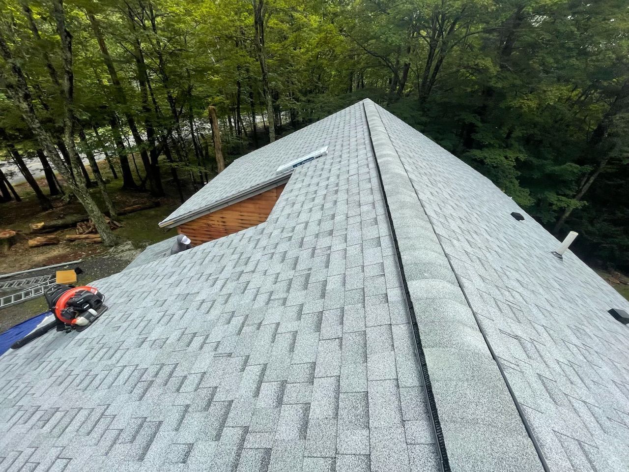 Roofing crew installing gray shingles on a steep roof beside a wooded area
