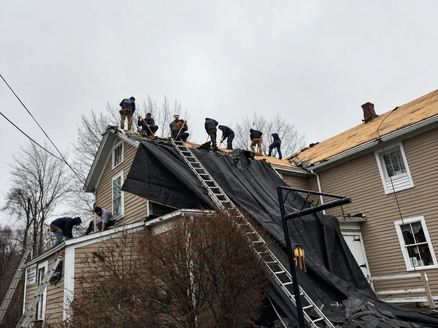 Workers on a house roof, removing a black tarp after storm damage, with ladders and debris visible.