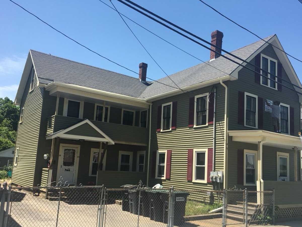 Two-story green house with a porch and chain-link fence under a clear blue sky
