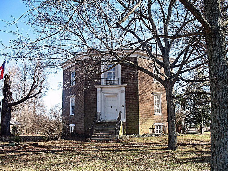 A large brick building with a white door is surrounded by trees and grass.