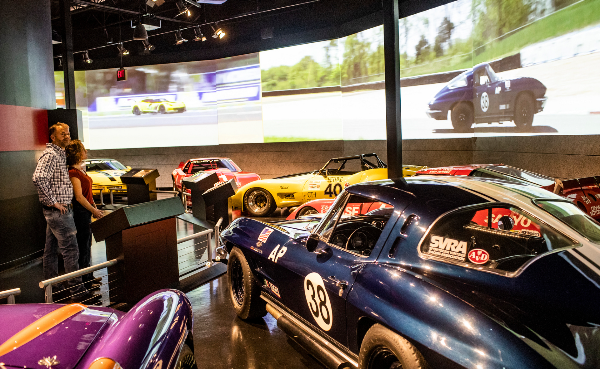 A group of cars are parked in a room with a large screen behind them.