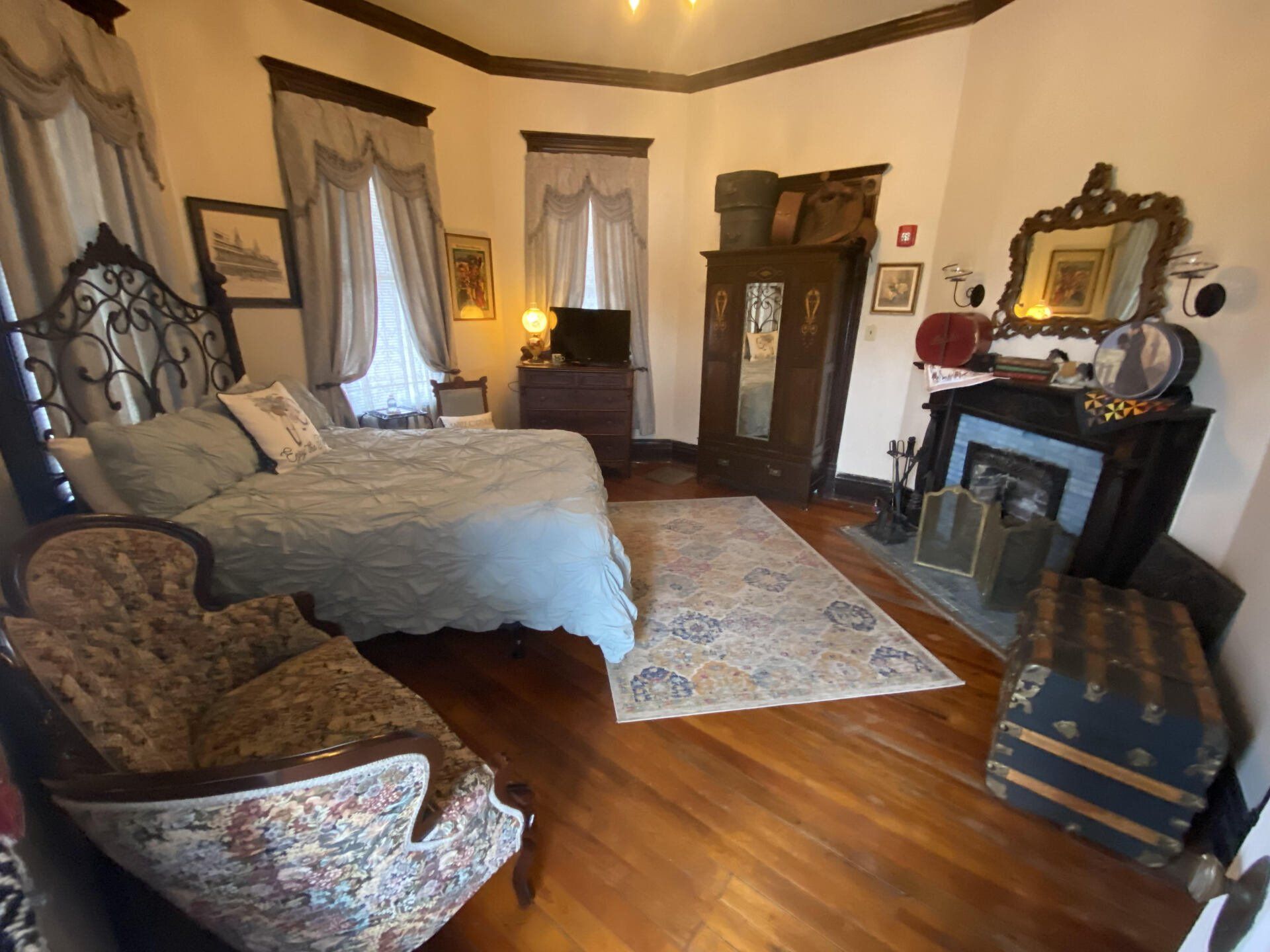A bedroom with a bed , chair , trunk and fireplace.