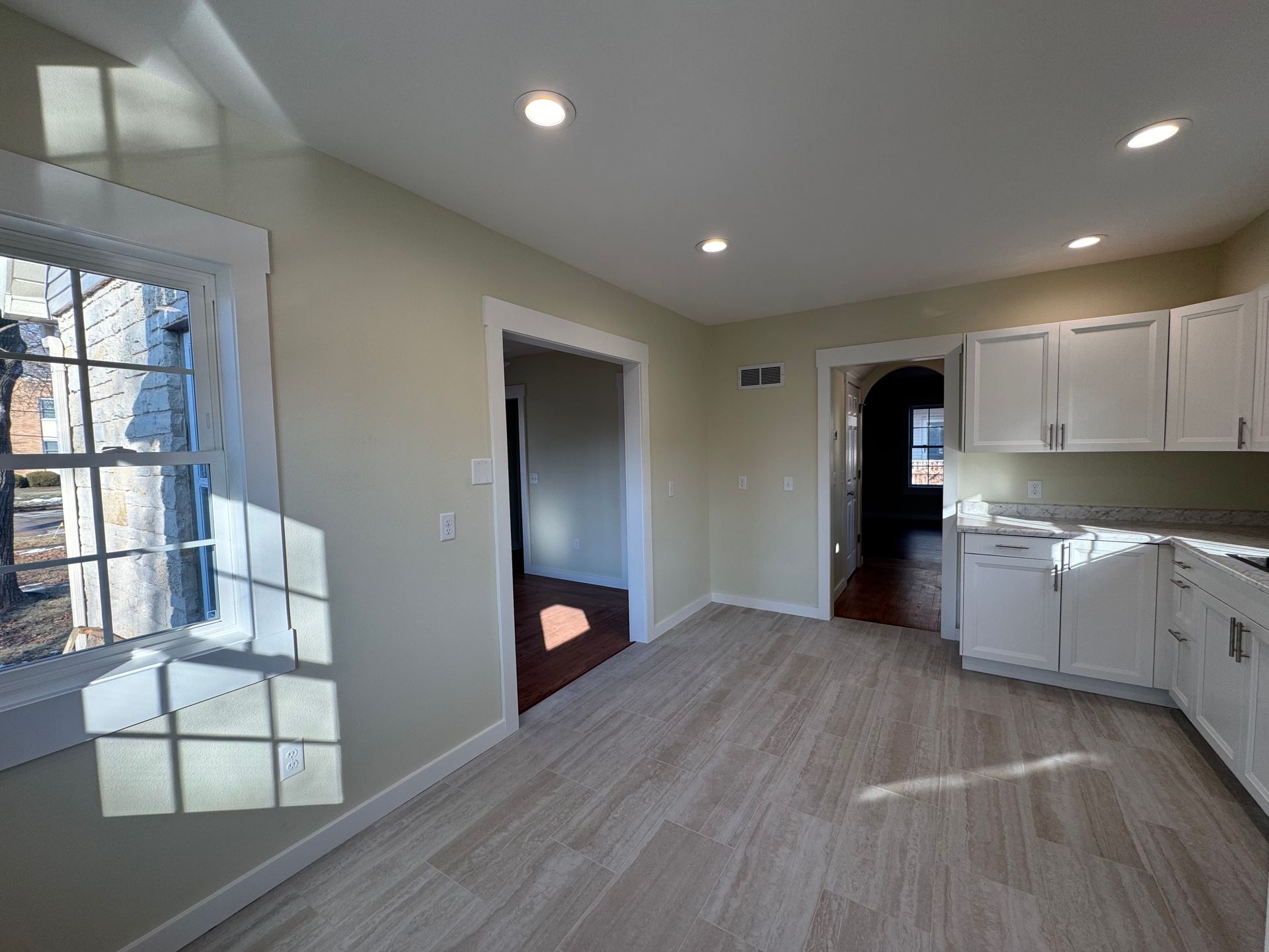 Bright kitchen with white cabinets, wood floors, recessed lights, and a large window by a doorway.
