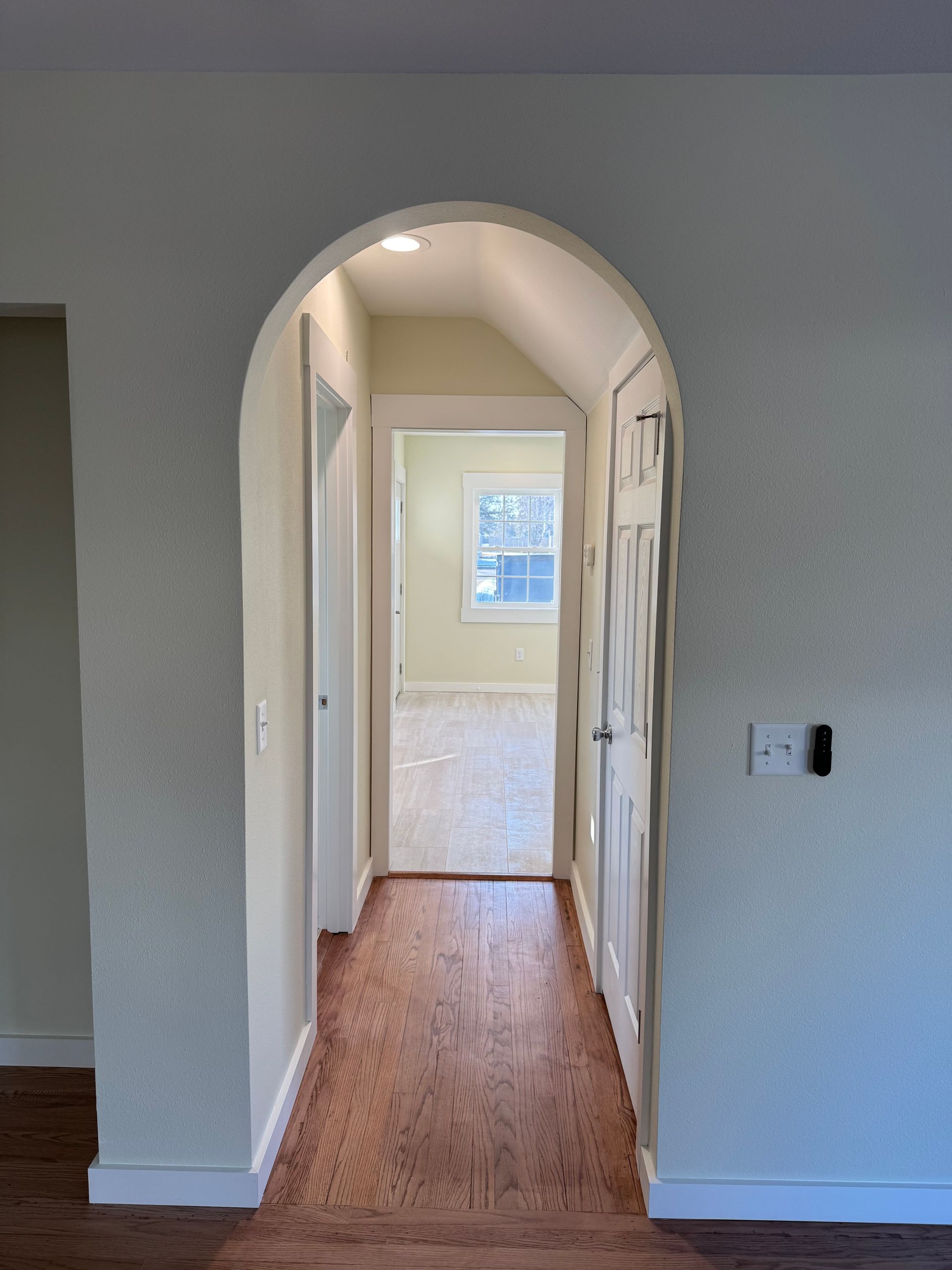 Arched hallway with wood floor, white walls, and a small window at the end