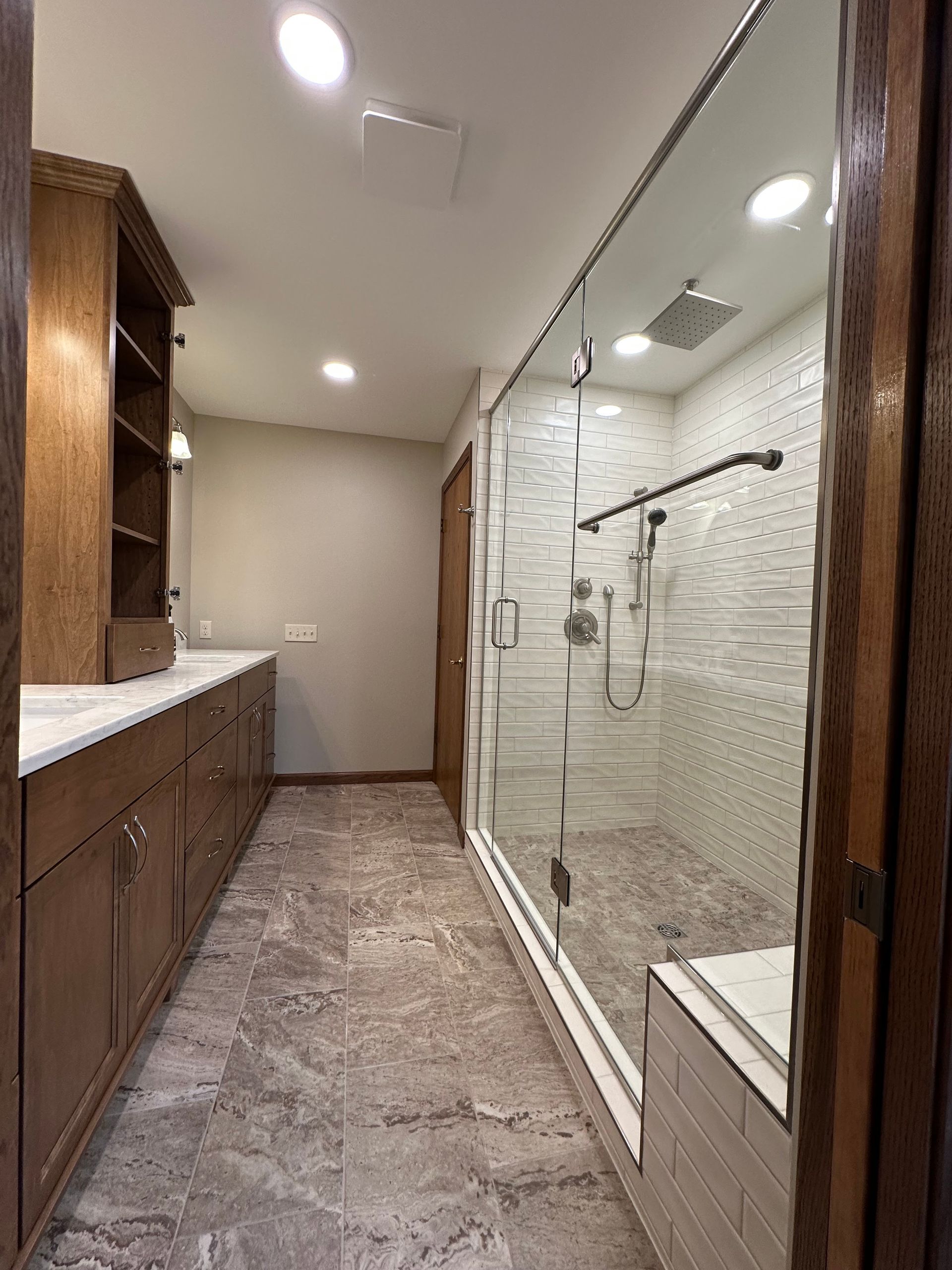 Long narrow bathroom with vanity on left and glass shower on right, beige tile and recessed lights
