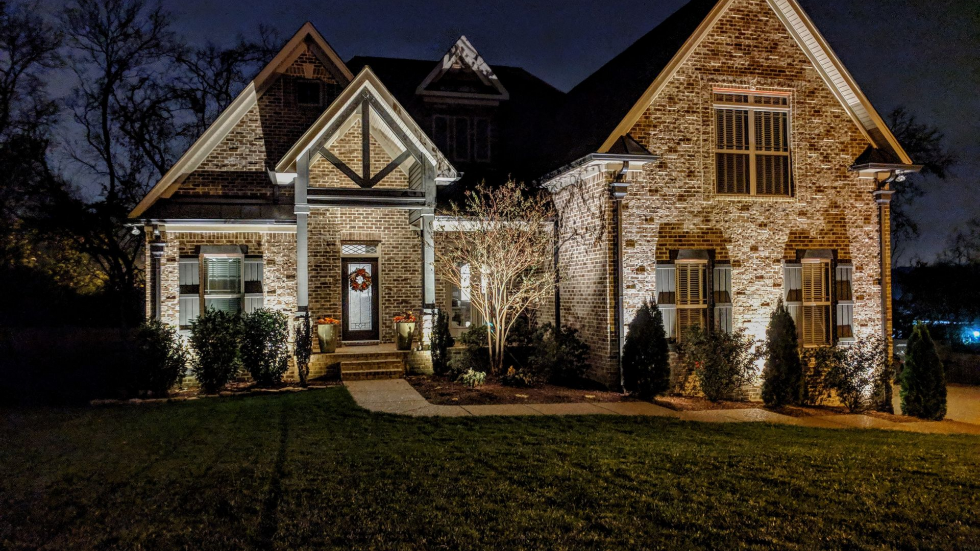 Brick suburban house lit warmly at night, with peaked gables and a dark lawn in front