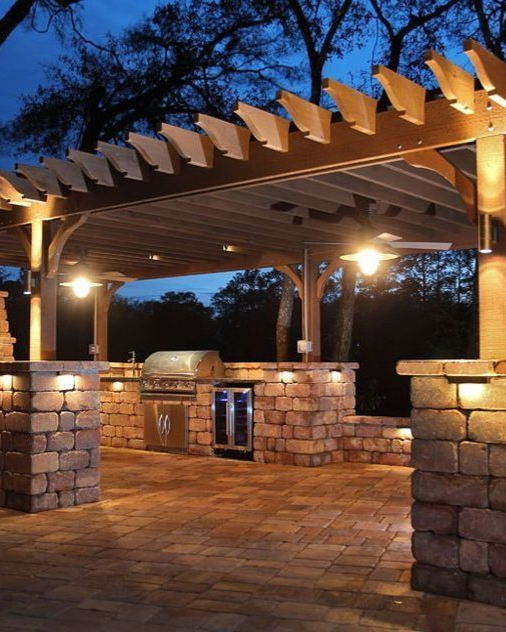 Stone patio pavilion with wooden pergola roof, warm lights, and an outdoor grill at dusk