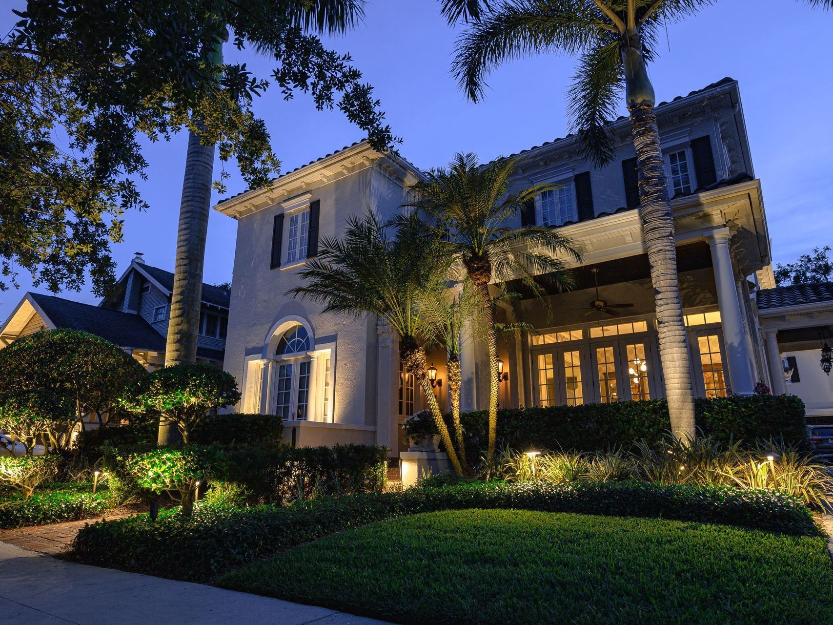 Large illuminated two-story house at dusk with palm trees and manicured landscaping