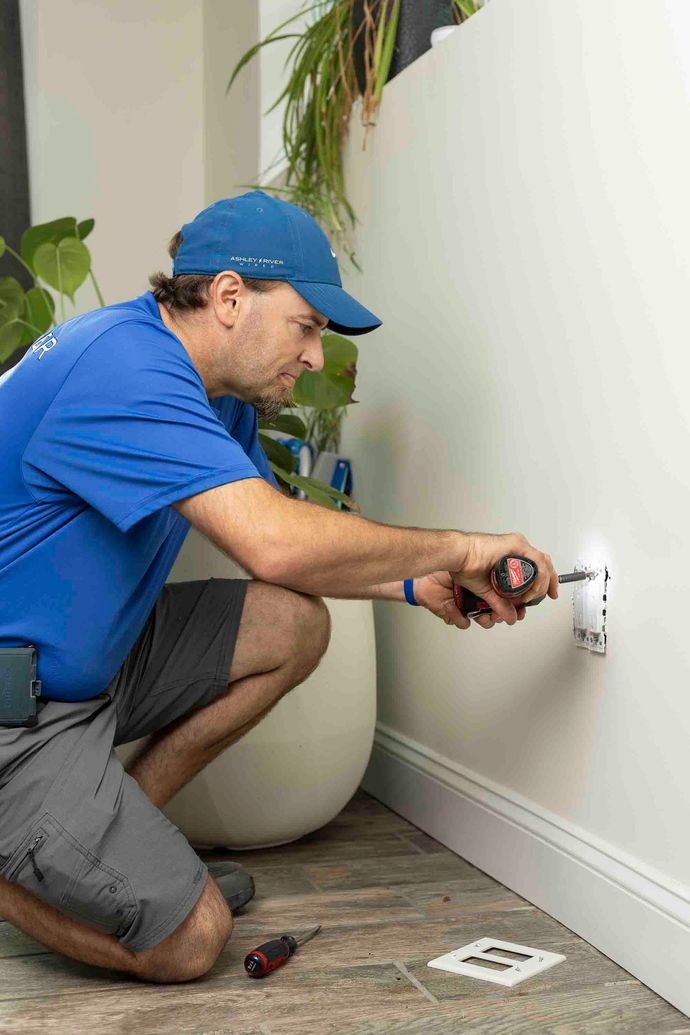 Electrician in blue uniform kneels by a wall outlet, using a screwdriver to install or repair it.