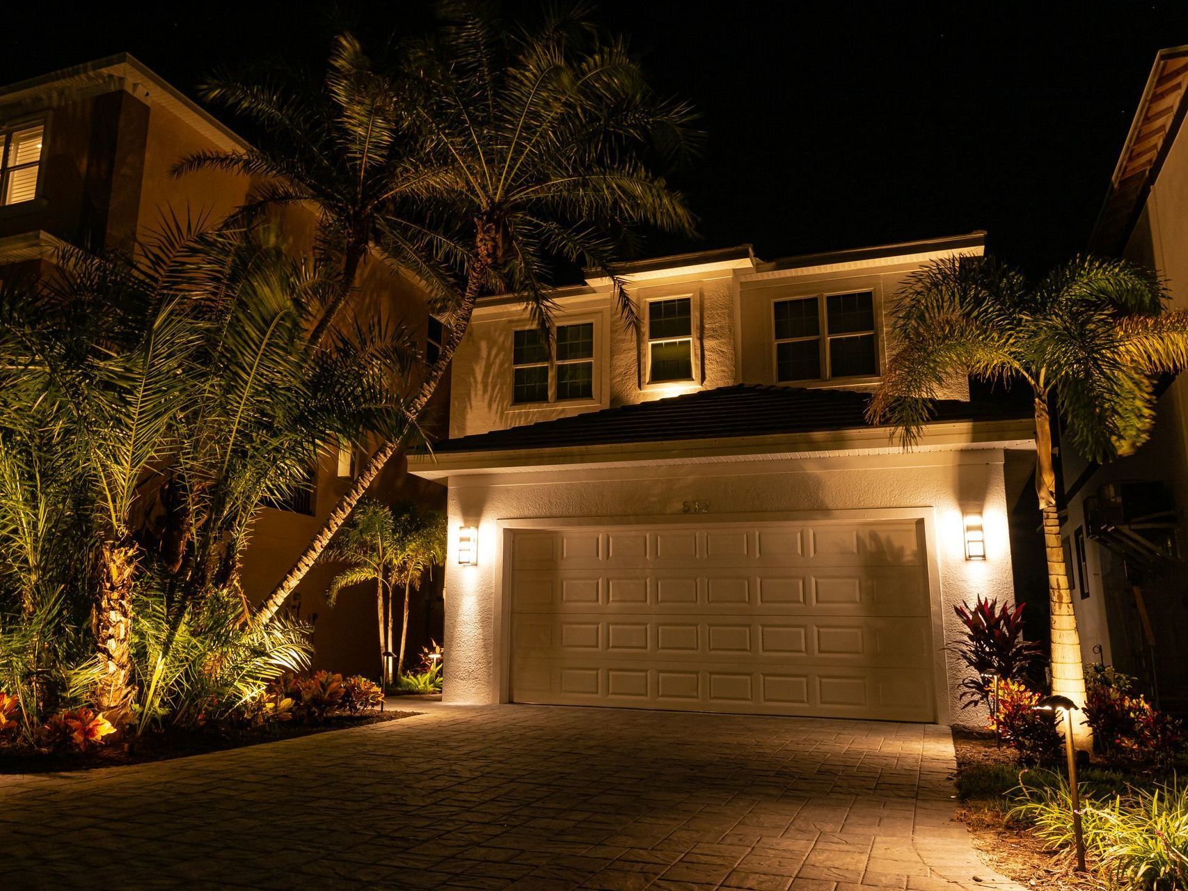 A warmly lit suburban house at night with palm trees and a two-car garage