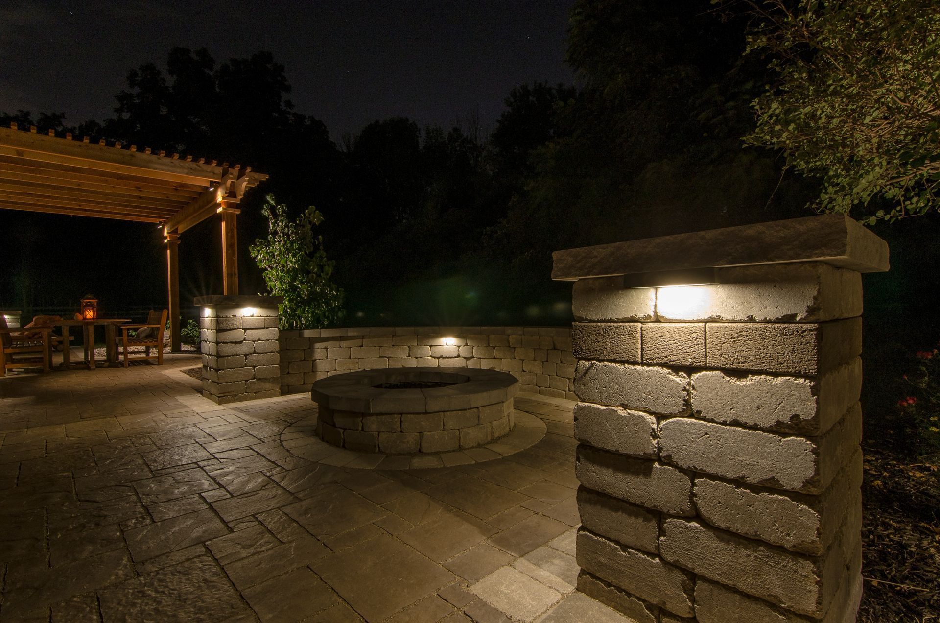 Nighttime stone patio with a lit fire pit and warm garden lights under a covered pergola