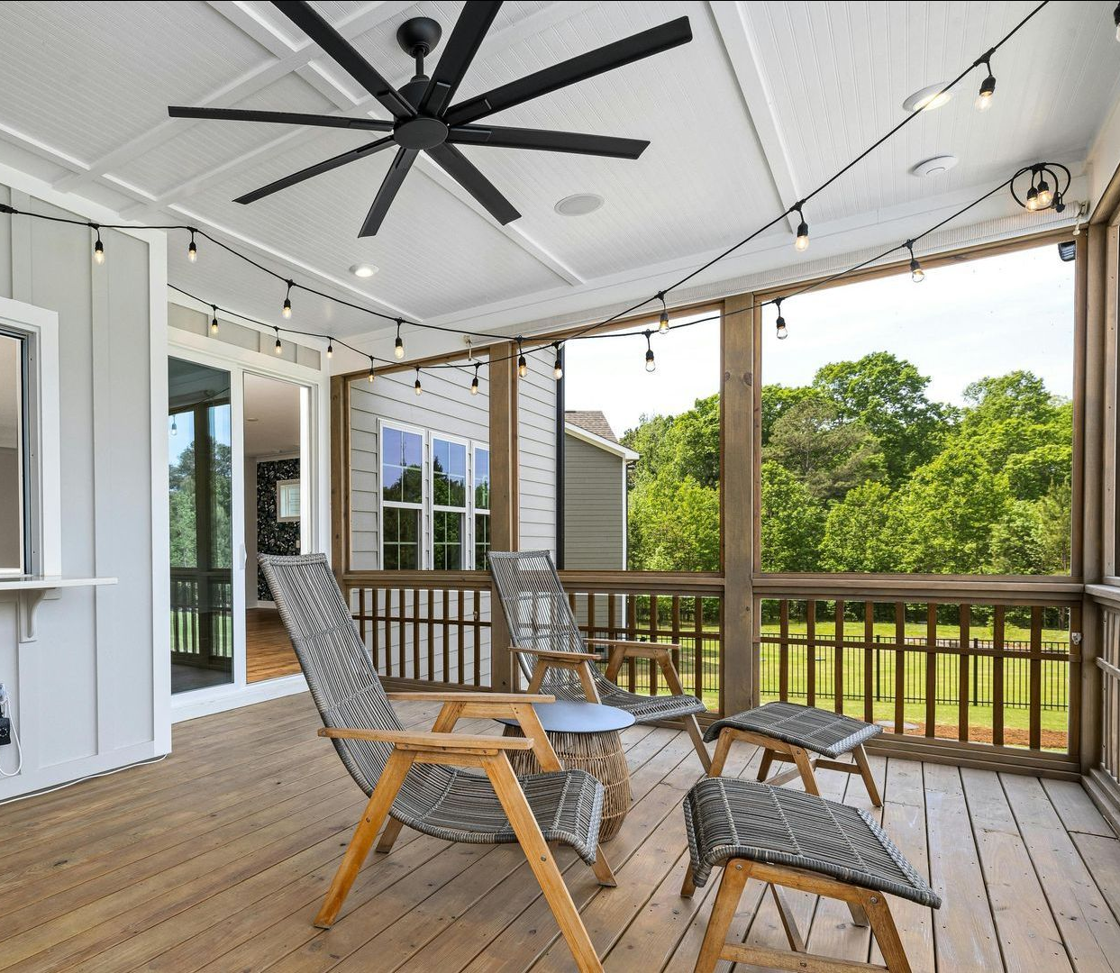Covered porch with wicker chairs, black ceiling fan, and a view of green trees and a yard.