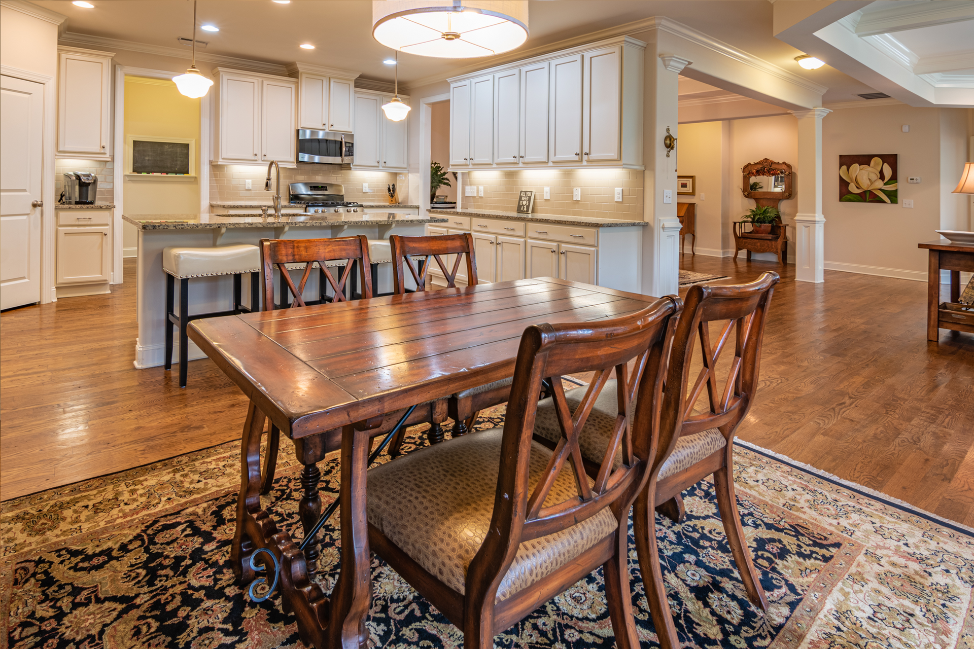 Bright dining room with wooden table, chairs, patterned rug, and open kitchen in the background