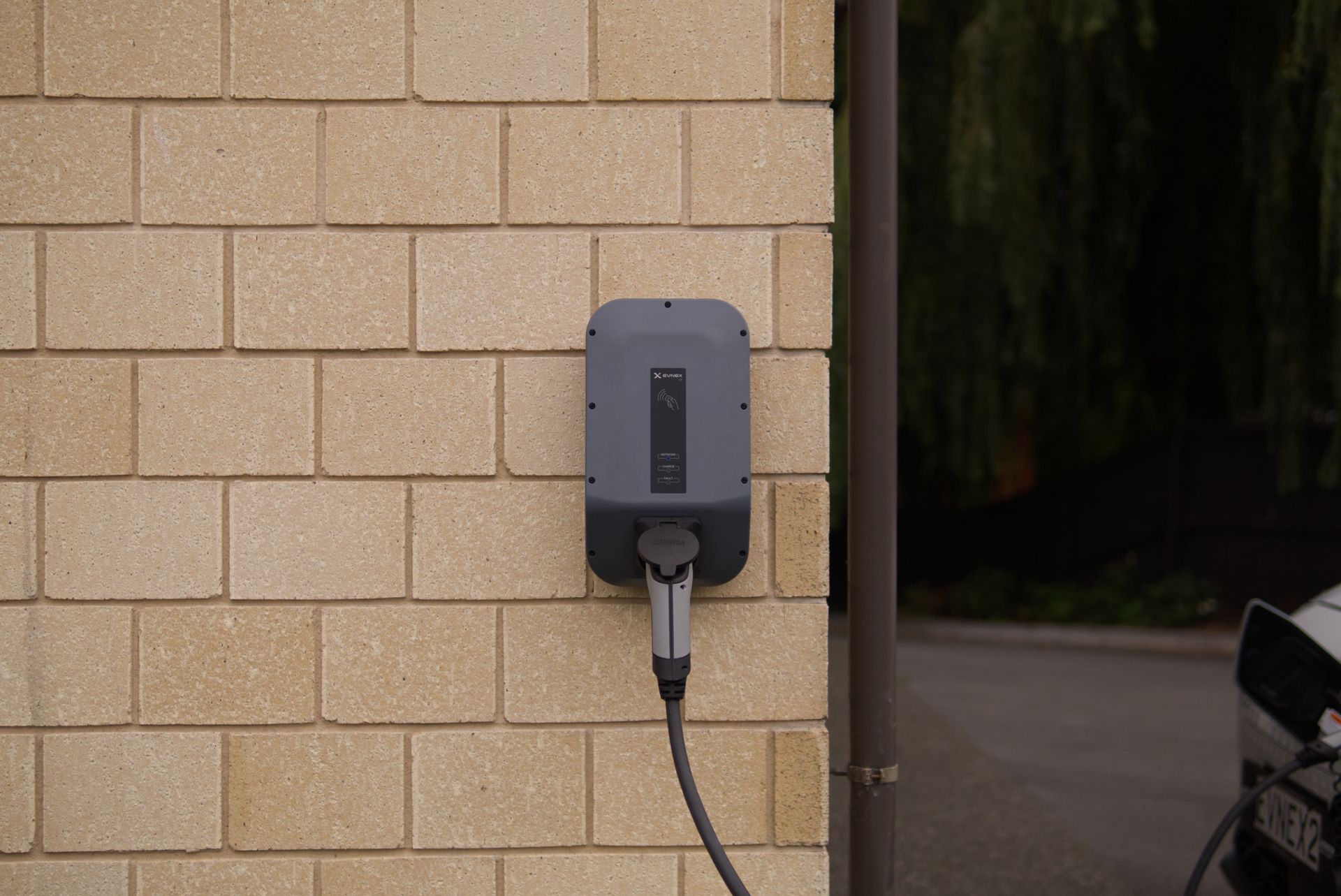 Wall-mounted EV charger on a beige brick exterior wall with a plugged-in cable