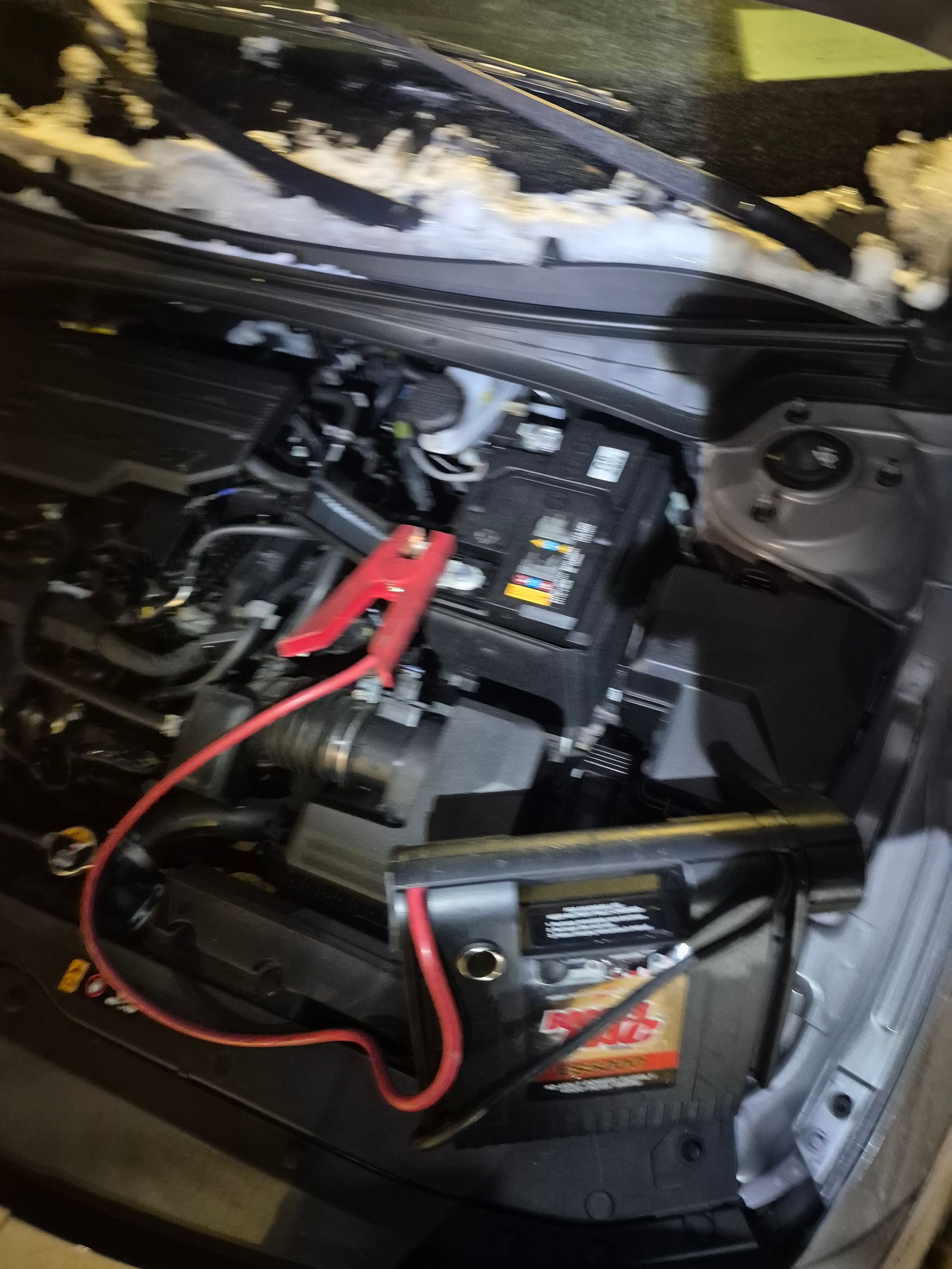Car battery in engine bay with red jumper cables attached to the terminals.