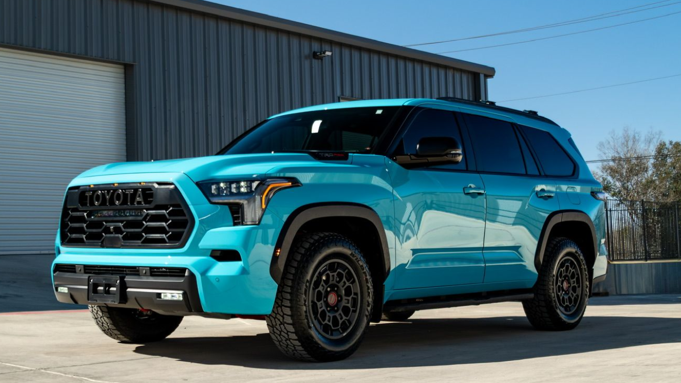 Rear side of a turquoise SUV with black off-road wheel and tire parked in sunlight.