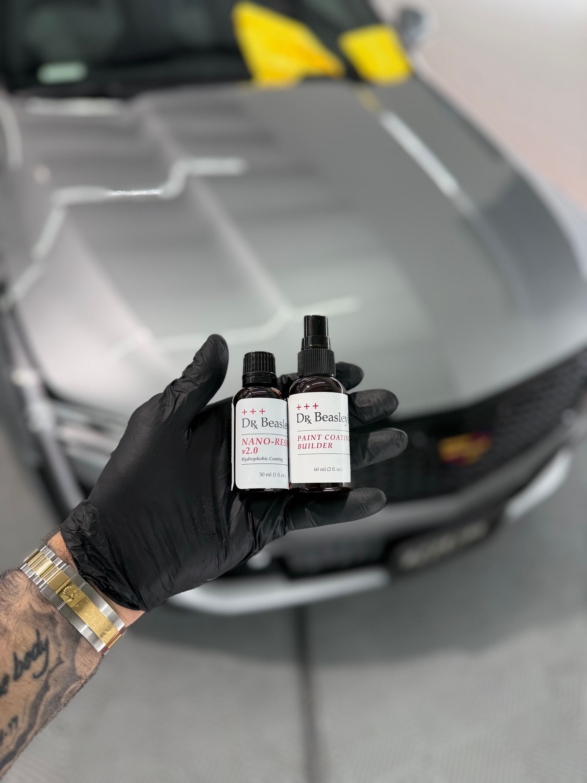 Gloved hand holding two small bottles in front of a gray sports car with yellow accents.