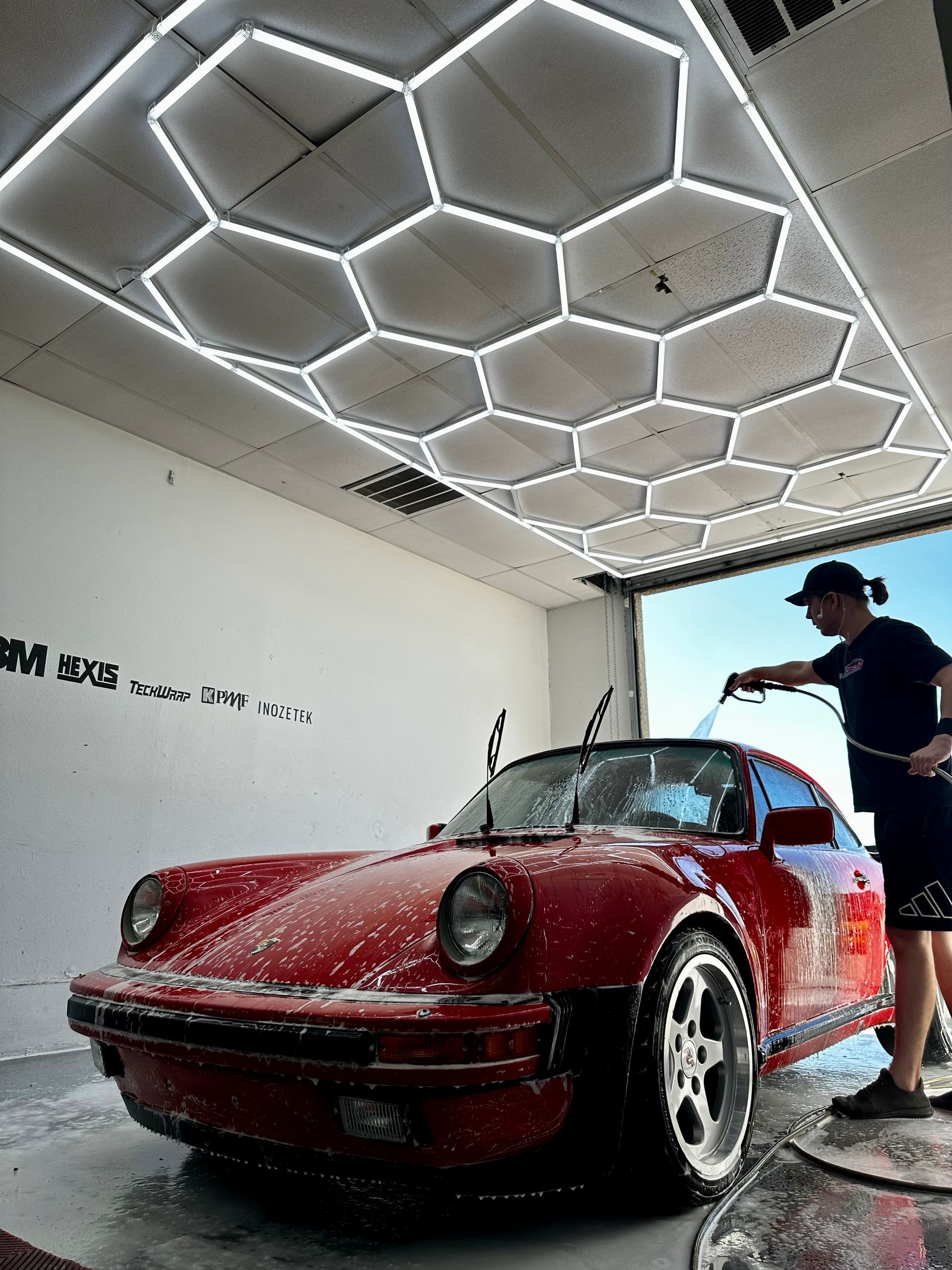 Red sports car being washed in a garage under hexagonal ceiling lights.