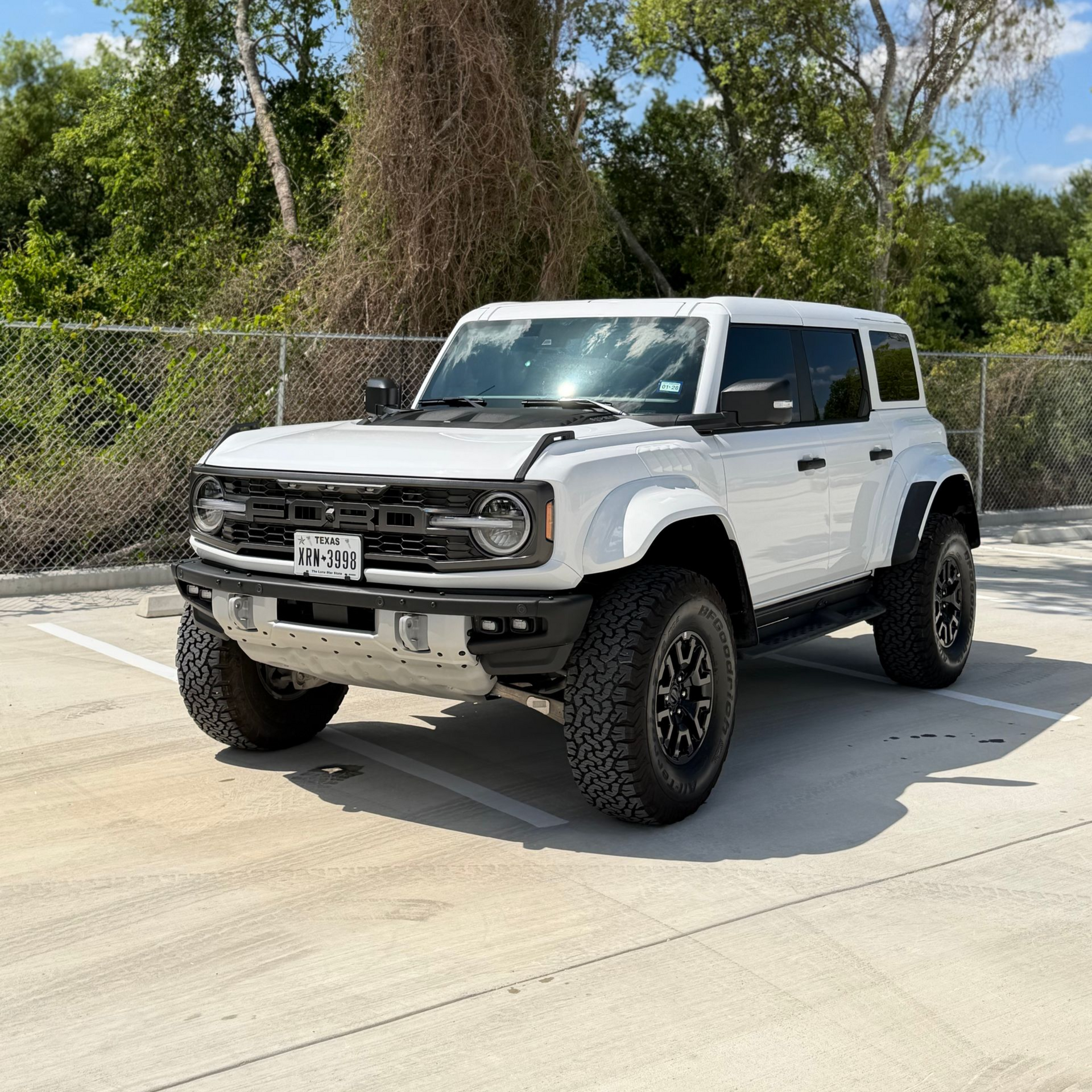 White Ford Bronco parked on a sunny concrete lot beside a chain-link fence and trees