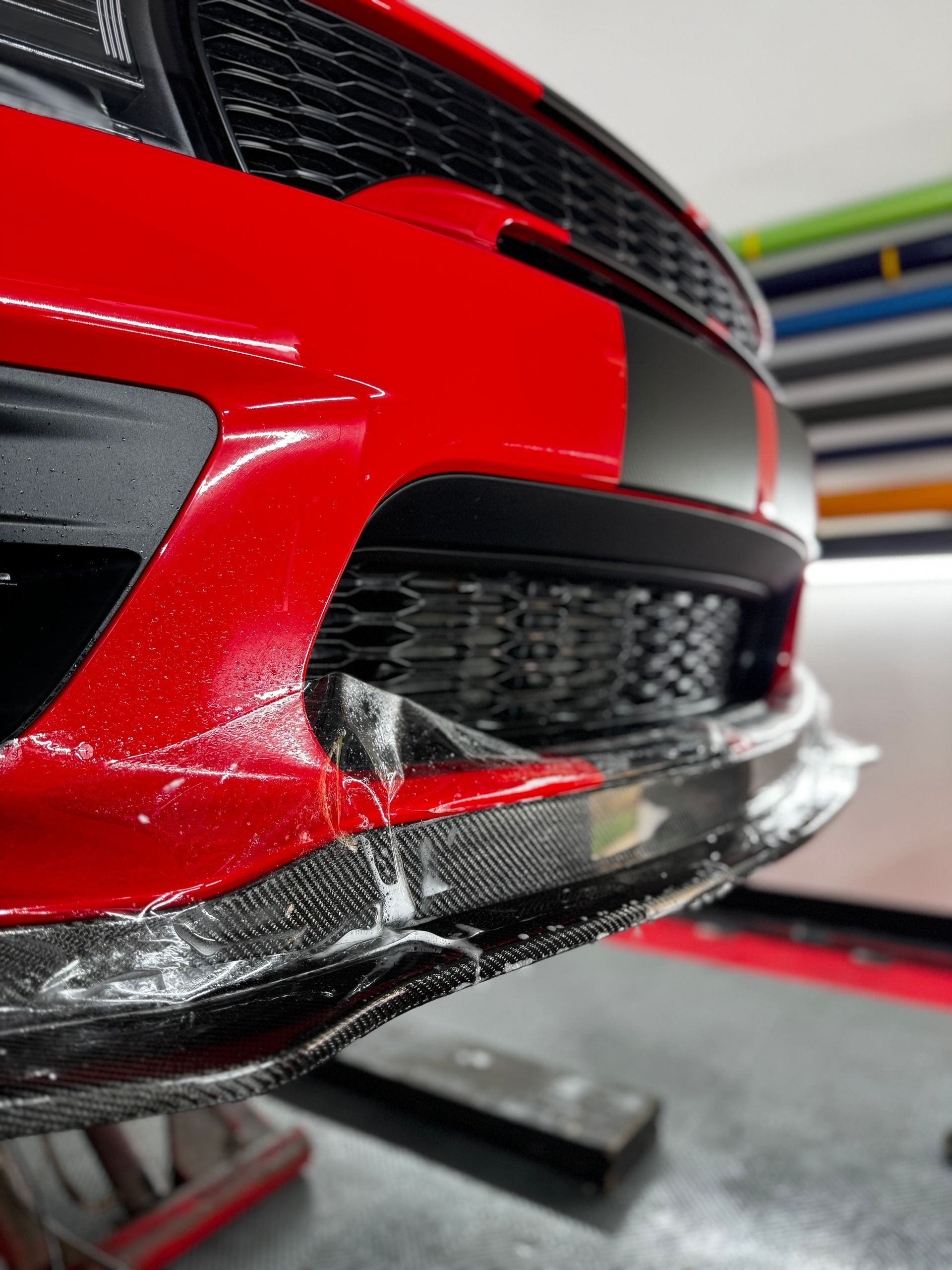 Close-up of a red sports car front bumper with black grille and carbon fiber splitter on a workshop floor