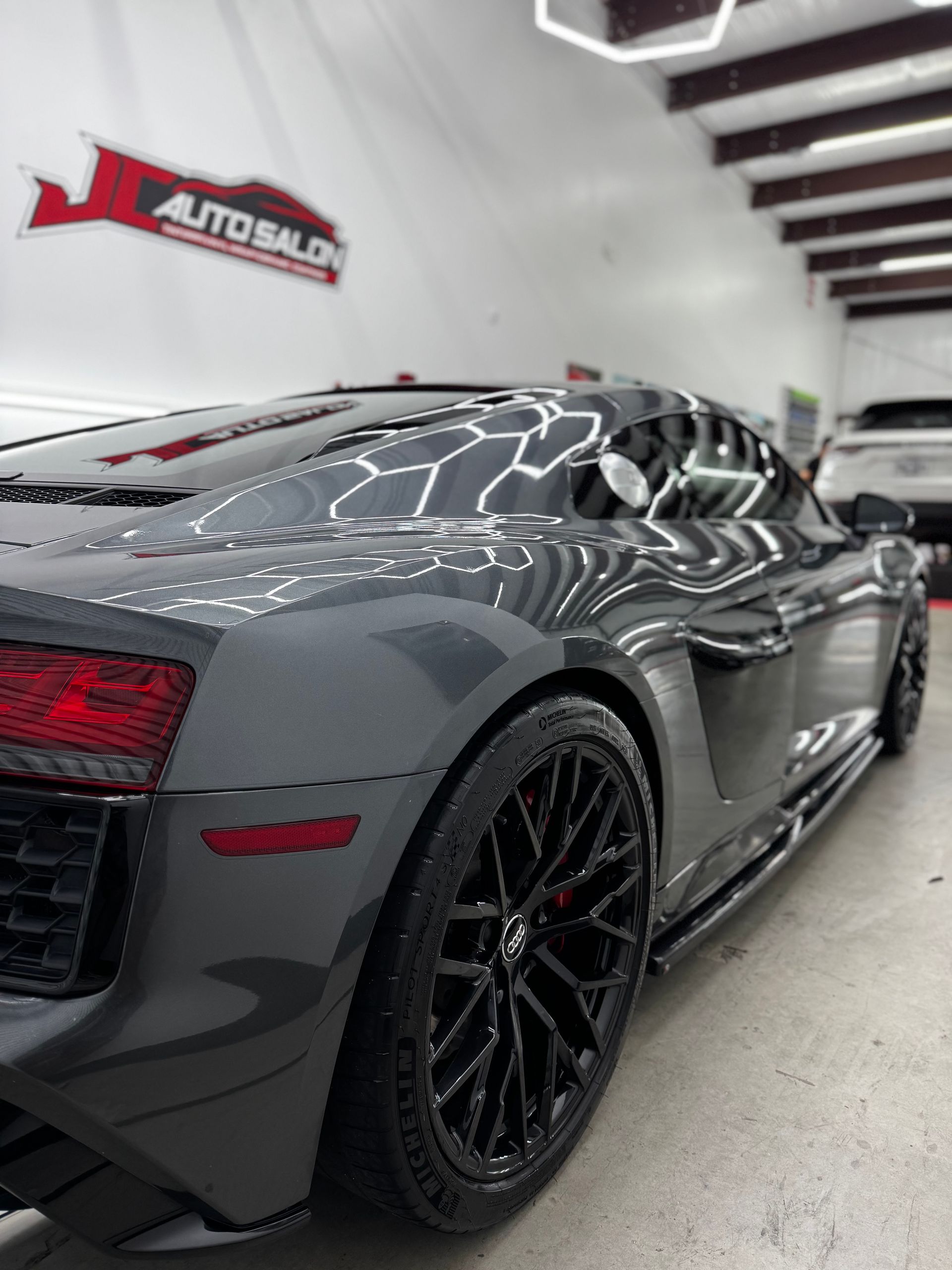 Glossy gray sports car in a showroom, rear side view, with black rims and red taillights.