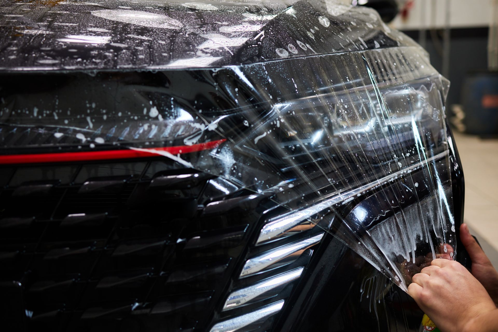 Hand wrapping clear protective film around a black car’s rear panel in a shop