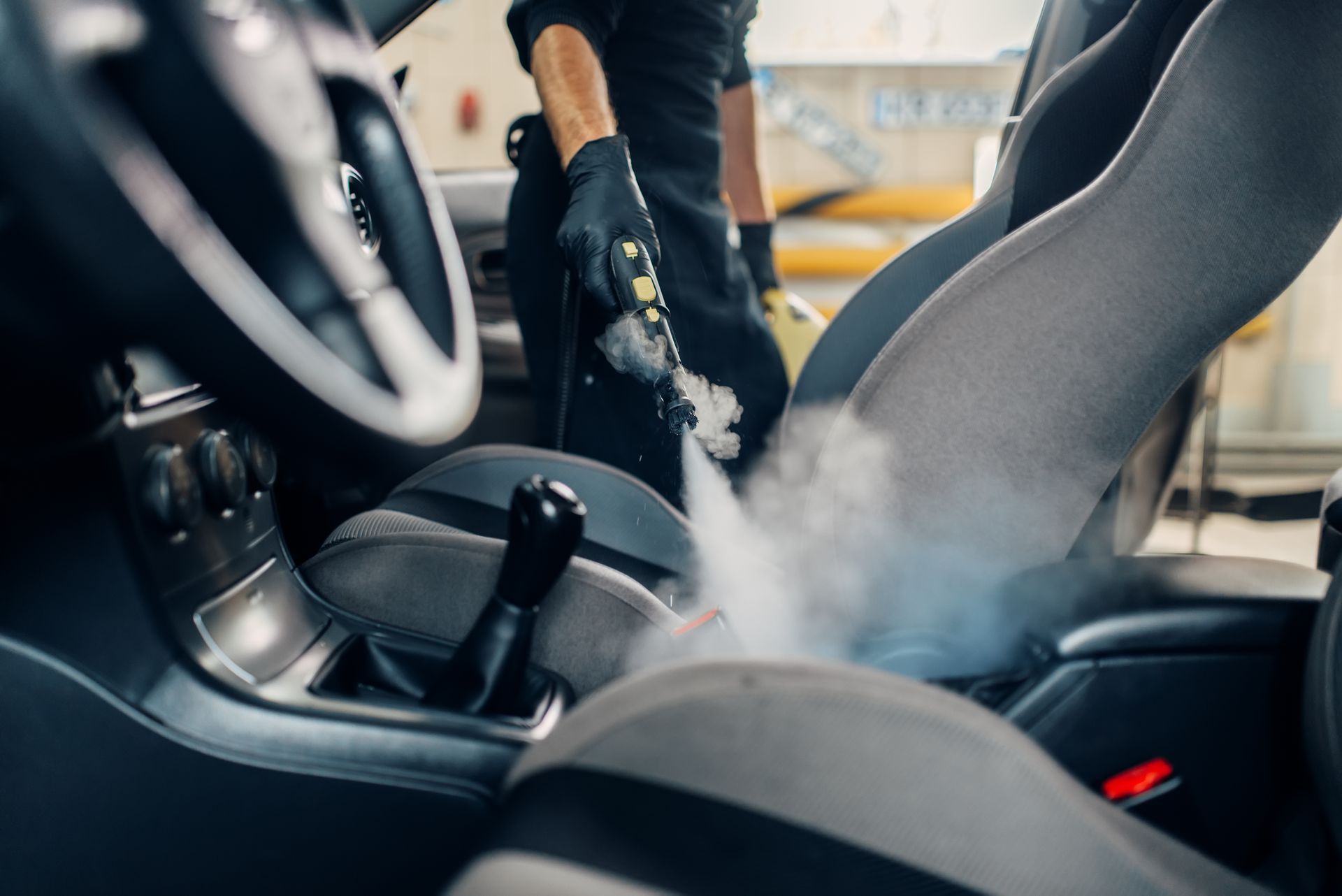 Car interior being steam cleaned, with a worker’s gloved hand spraying cleaner on the seats.