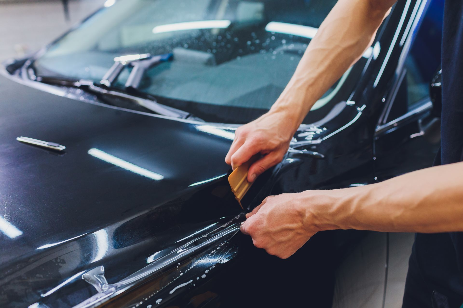 Hands applying protective film to a black car hood in a garage