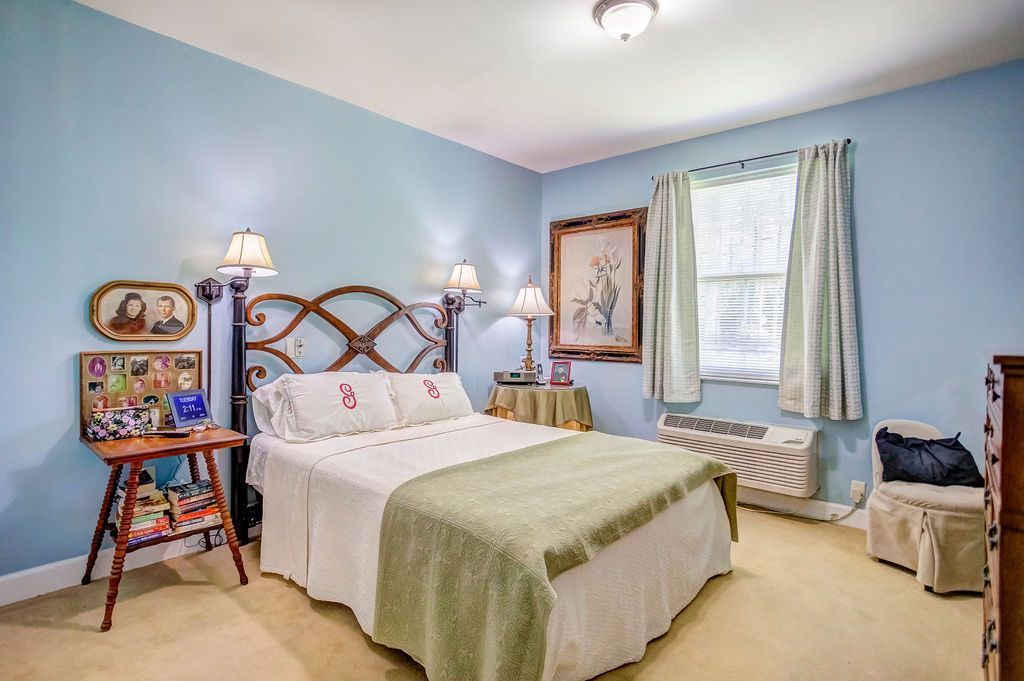 Light blue bedroom with a bed, nightstands, lamp, curtains, and a colorful easel by the window