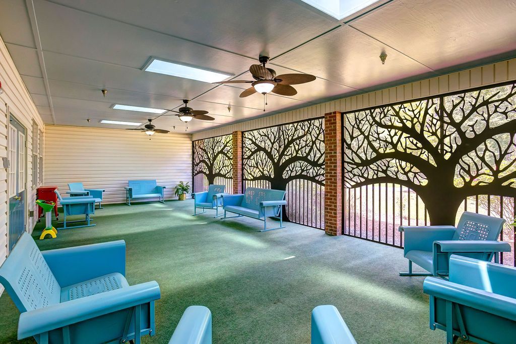 Covered patio with blue lounge chairs, green carpet, ceiling fans, and tree-patterned wall art