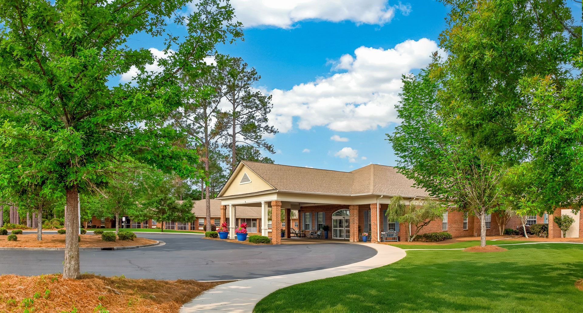 Brick clubhouse with covered entrance, surrounded by green trees and a curved driveway under a blue sky.