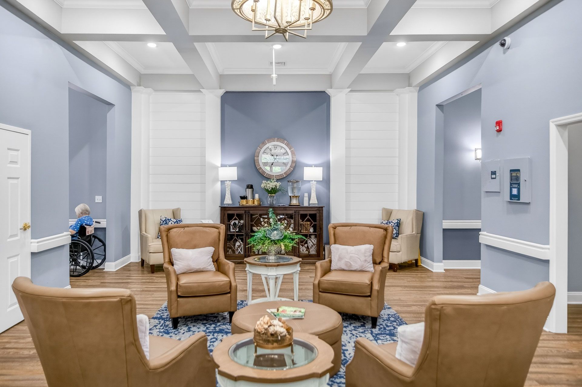 Elegant lobby seating area with tan armchairs, round tables, and blue-gray walls under chandeliers.